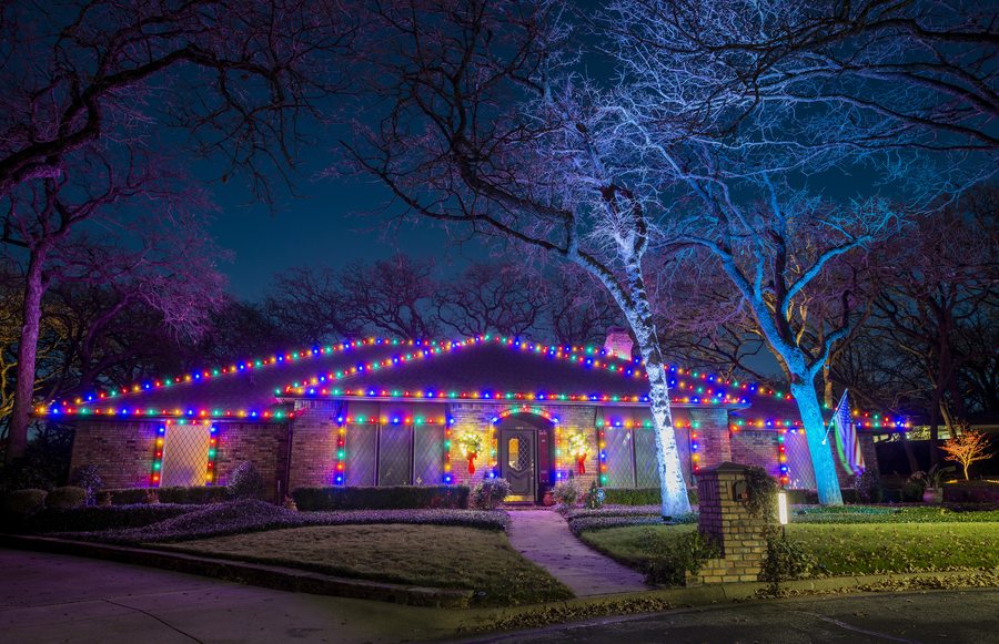 Brick ranch home outlined in multicolor Christmas lights with blue-lit tree at night