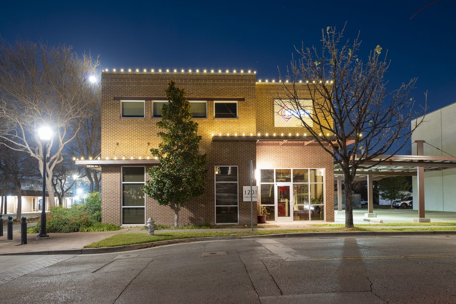Brick office building with warm white bistro-style holiday lights outlining the roofline and entrance at night