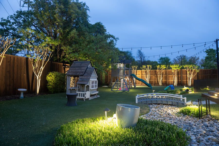 Playhouse and play structure lit across a turf backyard at dusk