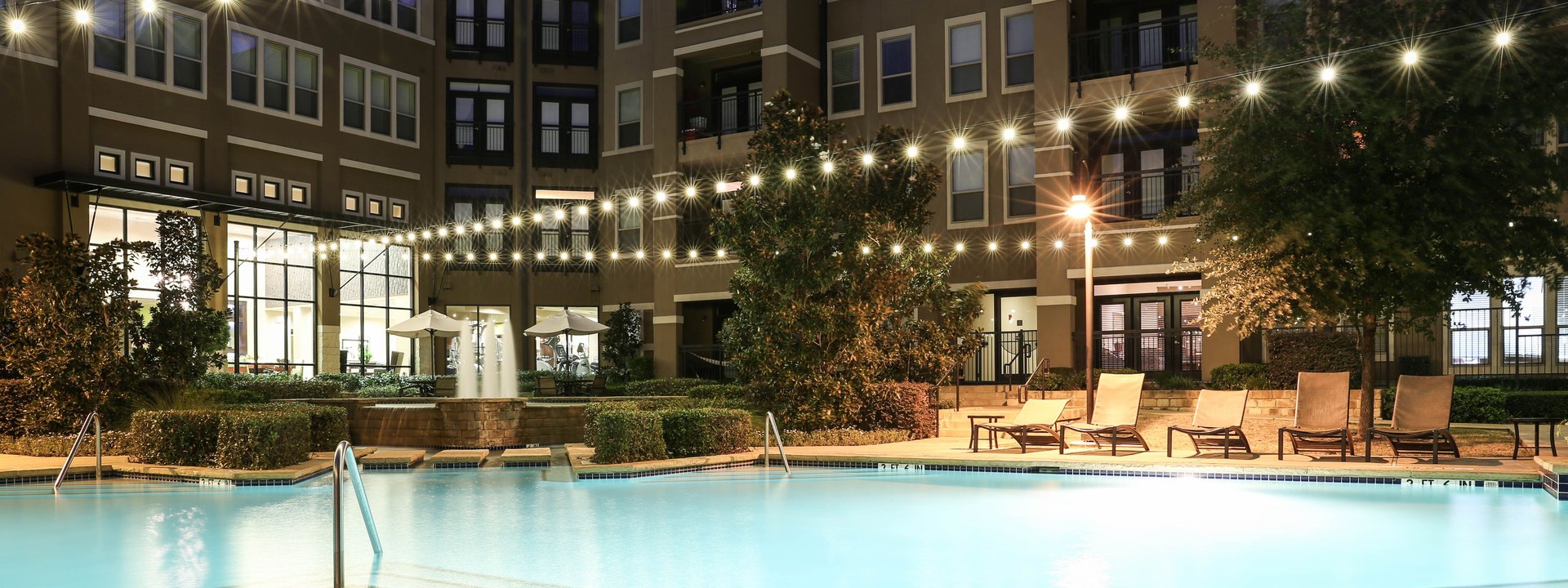 Apartment pool deck at night with string lights overhead reflecting in the water and lounge chairs along the edge